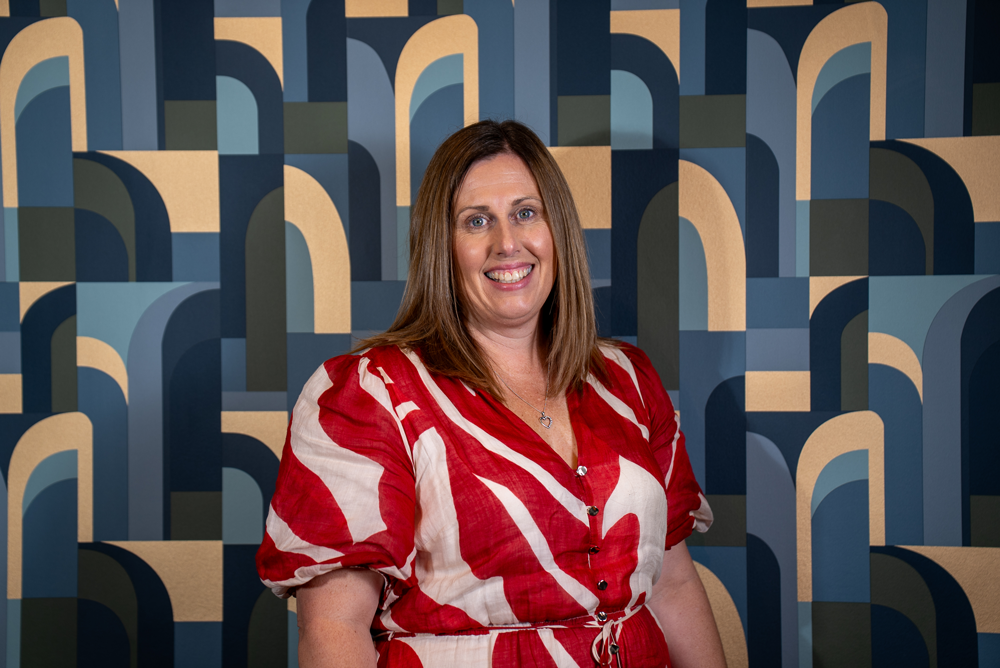 A woman with shoulder-length brown hair and a red and white patterned dress, smiling in front of a geometric blue, beige, and dark green painted backdrop.