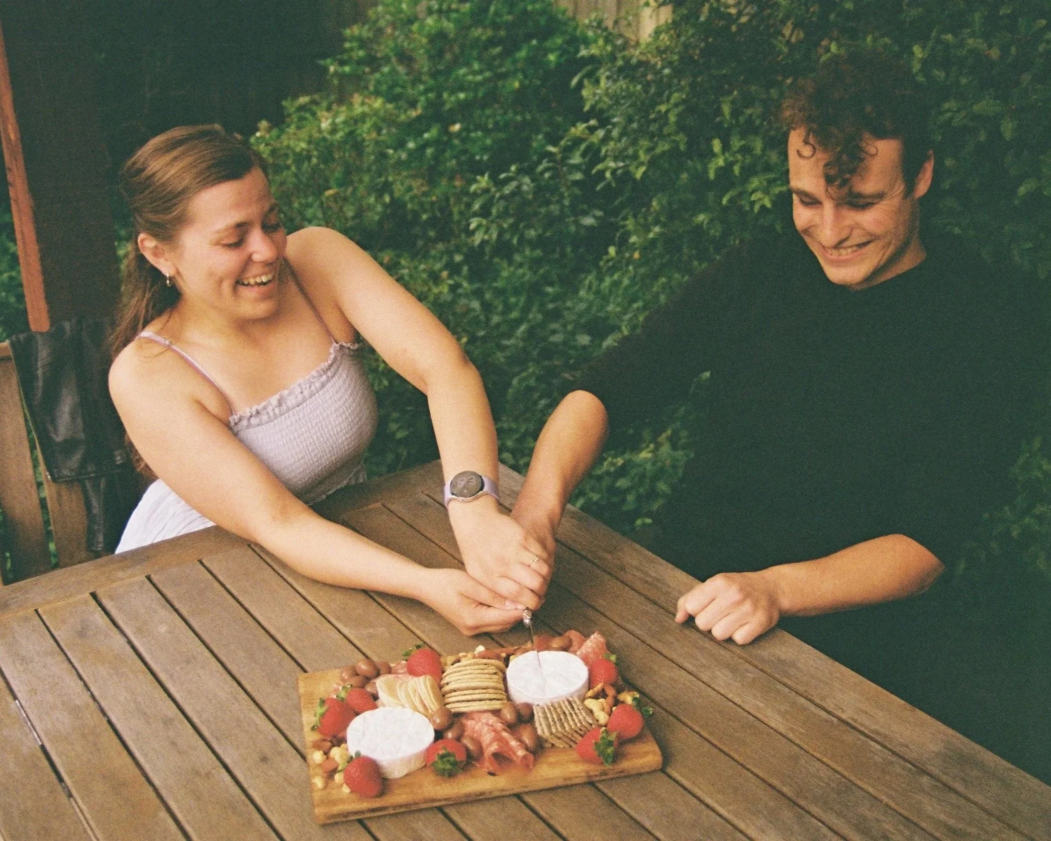 A woman and a man are sitting at a wooden table outdoors, smiling and holding a cheese knife together over a cheese platter with strawberries, cookies, crackers, and cured meats.