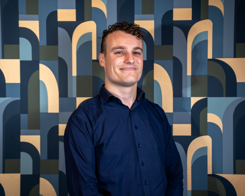 A man with short, curly dark hair, wearing a navy blue shirt, smiling slightly while standing in front of a modern, geometric-patterned background with arches and curves in shades of blue and beige.