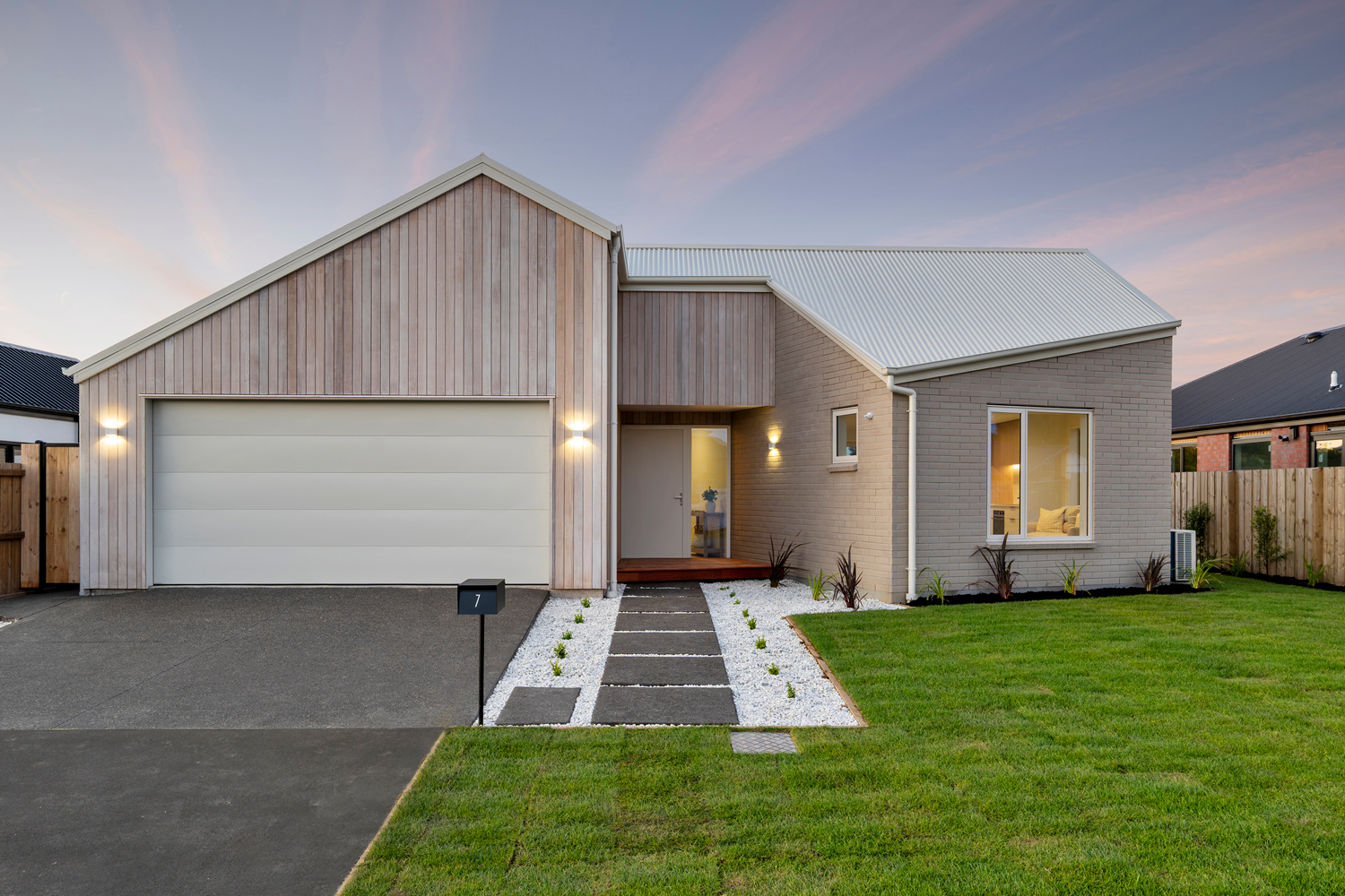 Modern suburban house with a light-colored brick and wood exterior, a white garage door, and a well-maintained front yard during sunset.