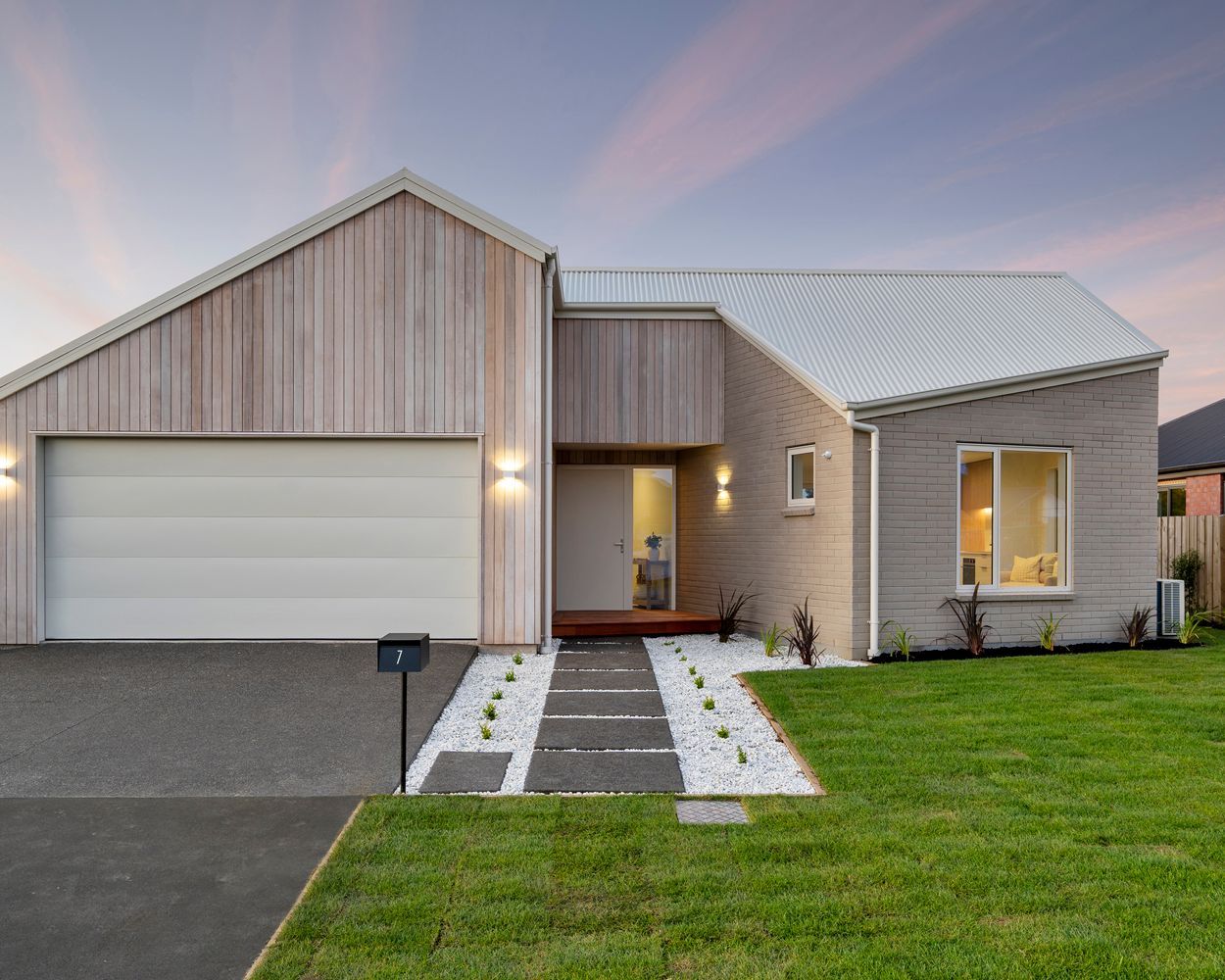 Modern house with brick and wood exterior, white garage door, manicured lawn, pathway, and sunset sky.