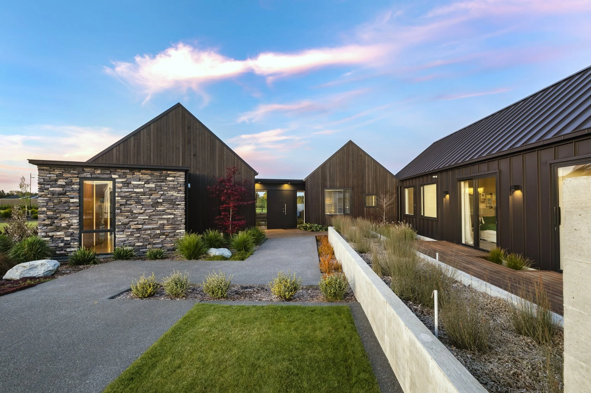 Modern house with stone and dark wood exterior, landscaped yard, pathway, and a wooden deck at sunset.