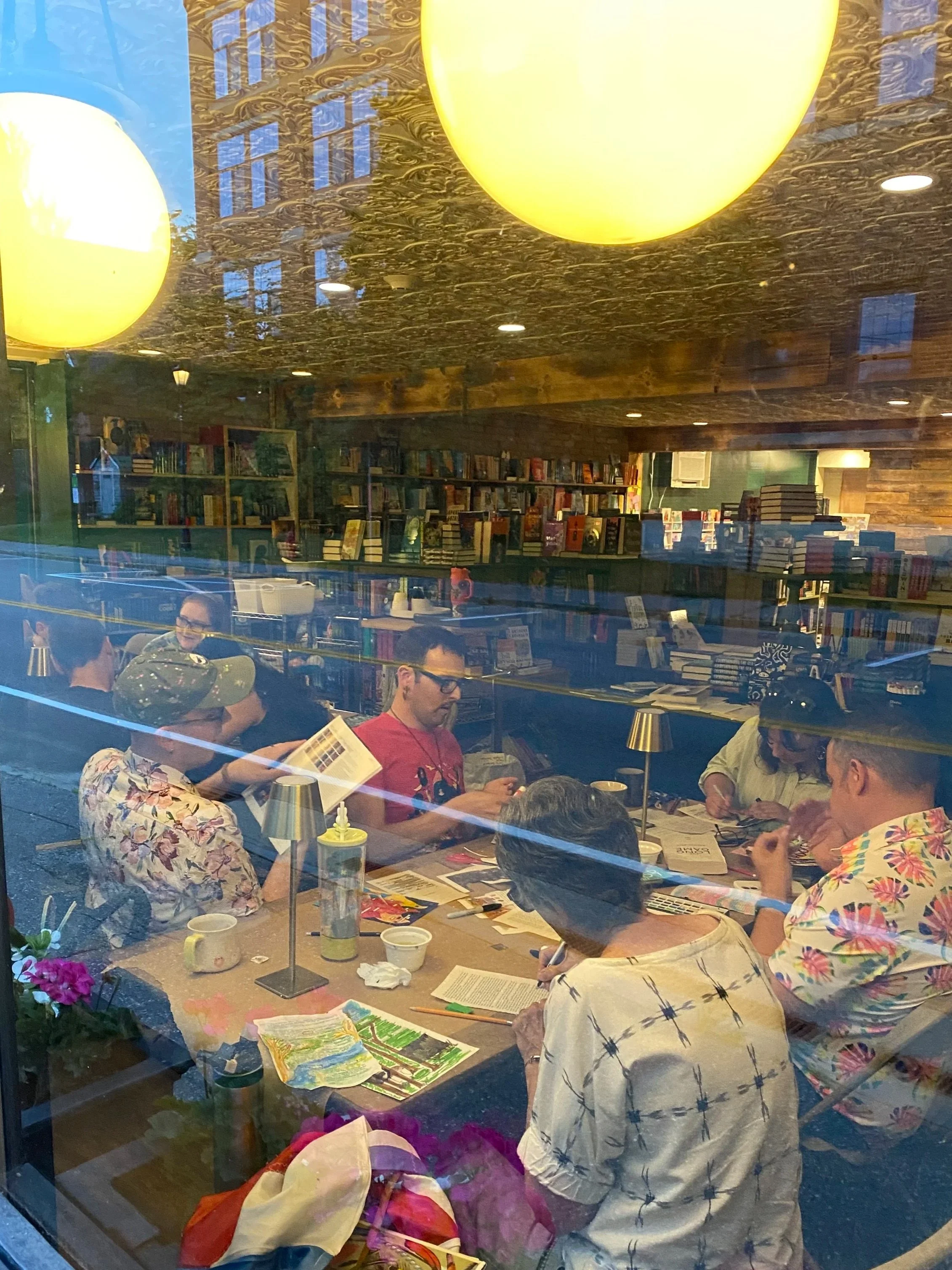 People sitting around a table inside a bookstore, reading and discussing. Bookshelves filled with books are visible in the background. The scene is viewed through a window reflecting the outside buildings.
