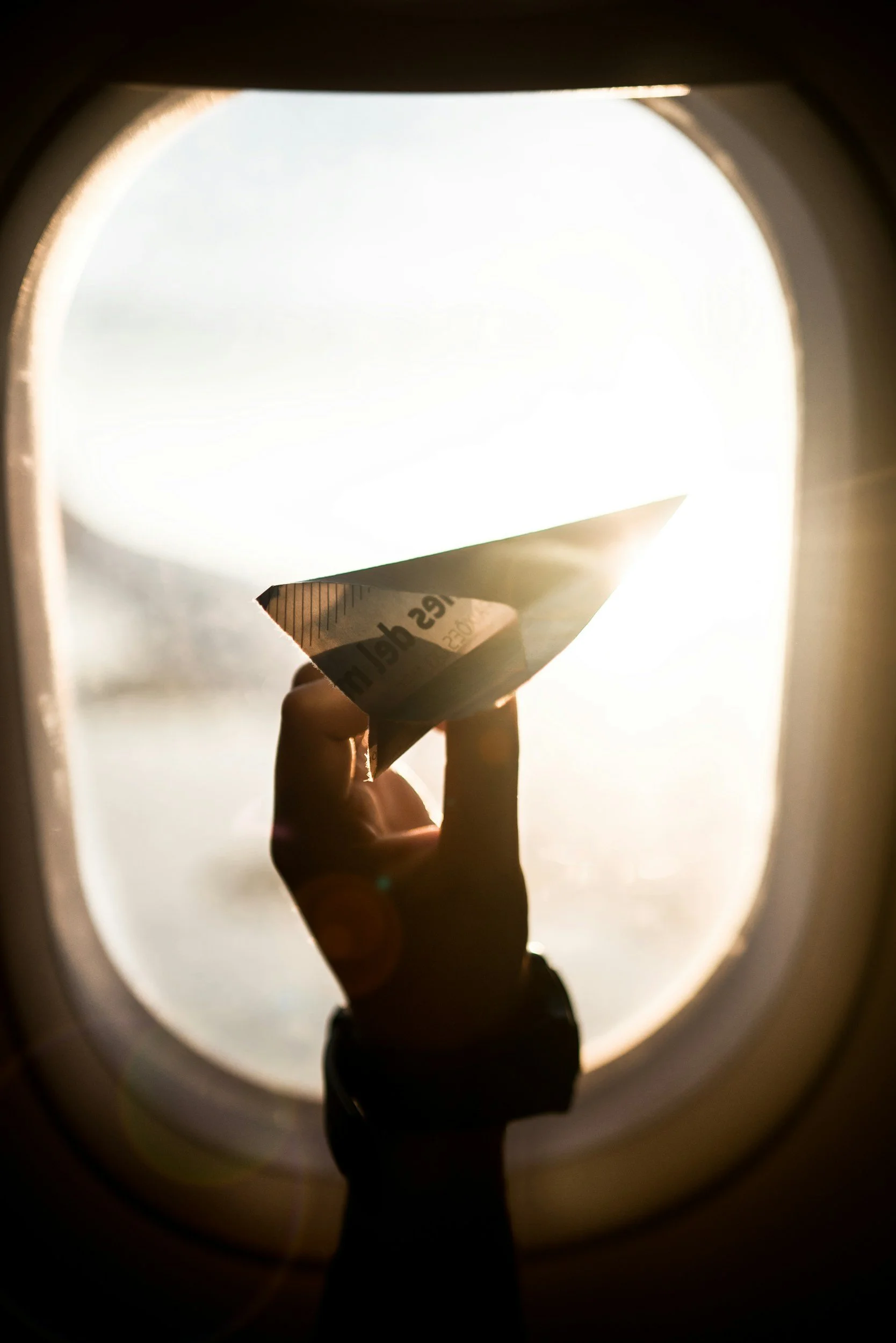 A hand holding a paper airplane inside an airplane window with sunlight shining outside.
