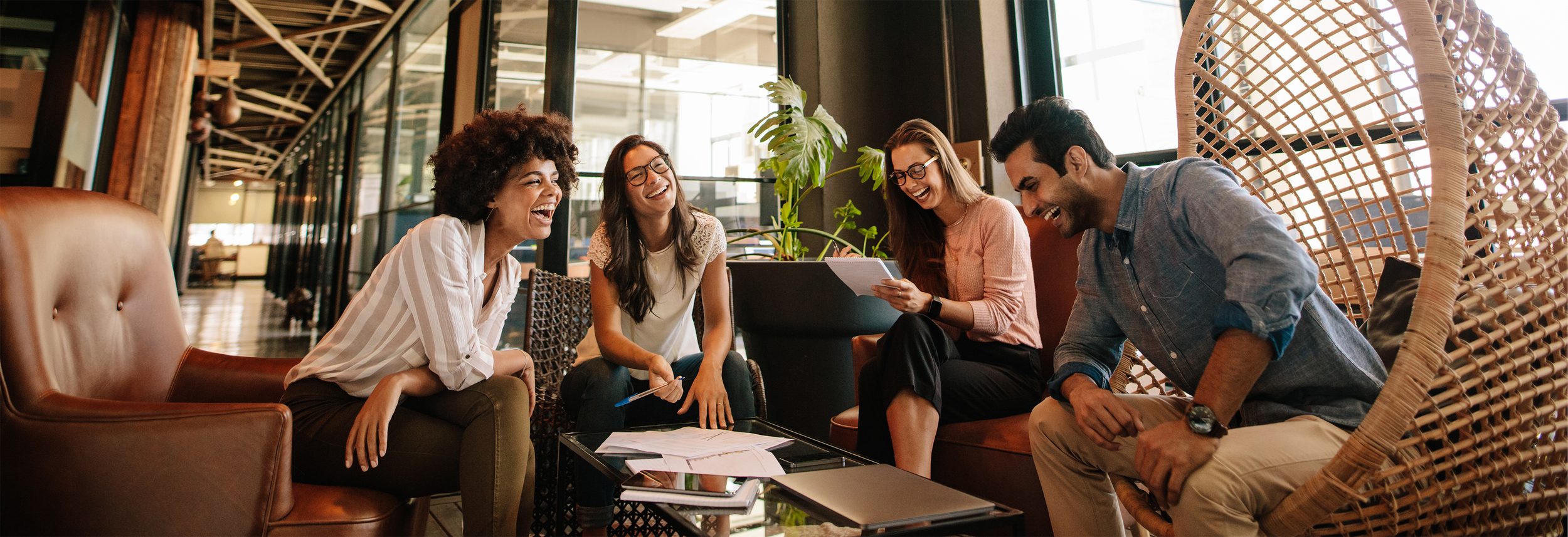 Group of five young adults sitting and laughing around a coffee table in a modern, well-lit café or lounge area, engaging in conversation and sharing a moment of joy.