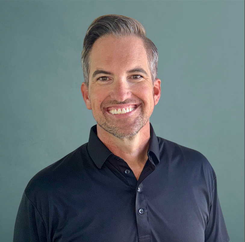 Smiling middle-aged man with short gray hair, wearing a black collared shirt, standing against a plain gray background.