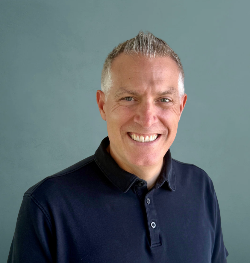Portrait of a smiling man with short, styled gray hair wearing a black collared shirt against a plain blue-green background.