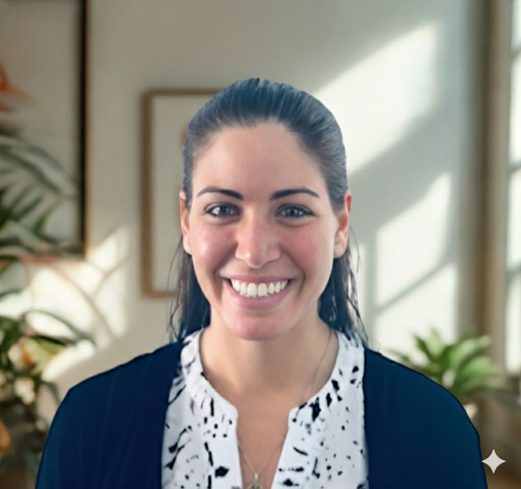 A woman with dark, pulled-back hair and blue eyes smiling, wearing a white blouse with black spots and a black cardigan, standing in a well-lit room with plants and framed art in the background.