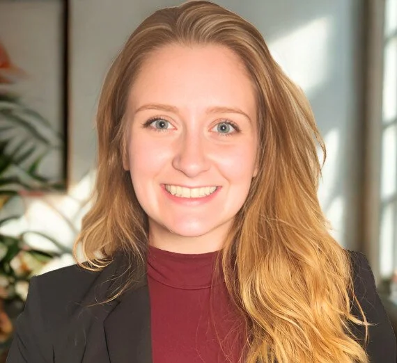 A woman with long red hair, blue eyes, and a bright smile, wearing a black blazer and a maroon turtleneck, in a well-lit indoor setting with plants in the background.
