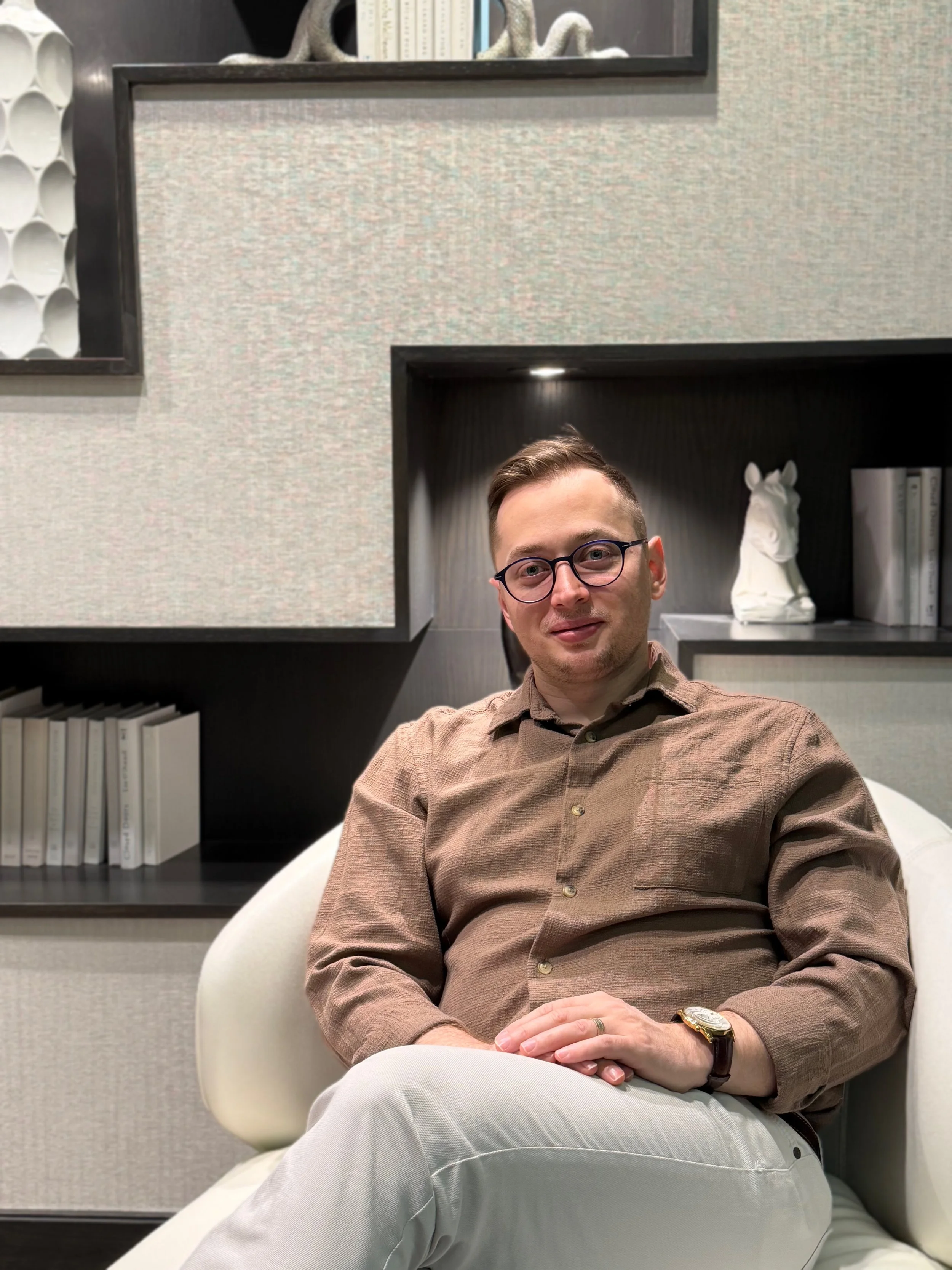 Kadir Iskesen with glasses and a wristwatch sitting in a modern chair in front of a bookshelf wall, with books and decorative horse sculptures.