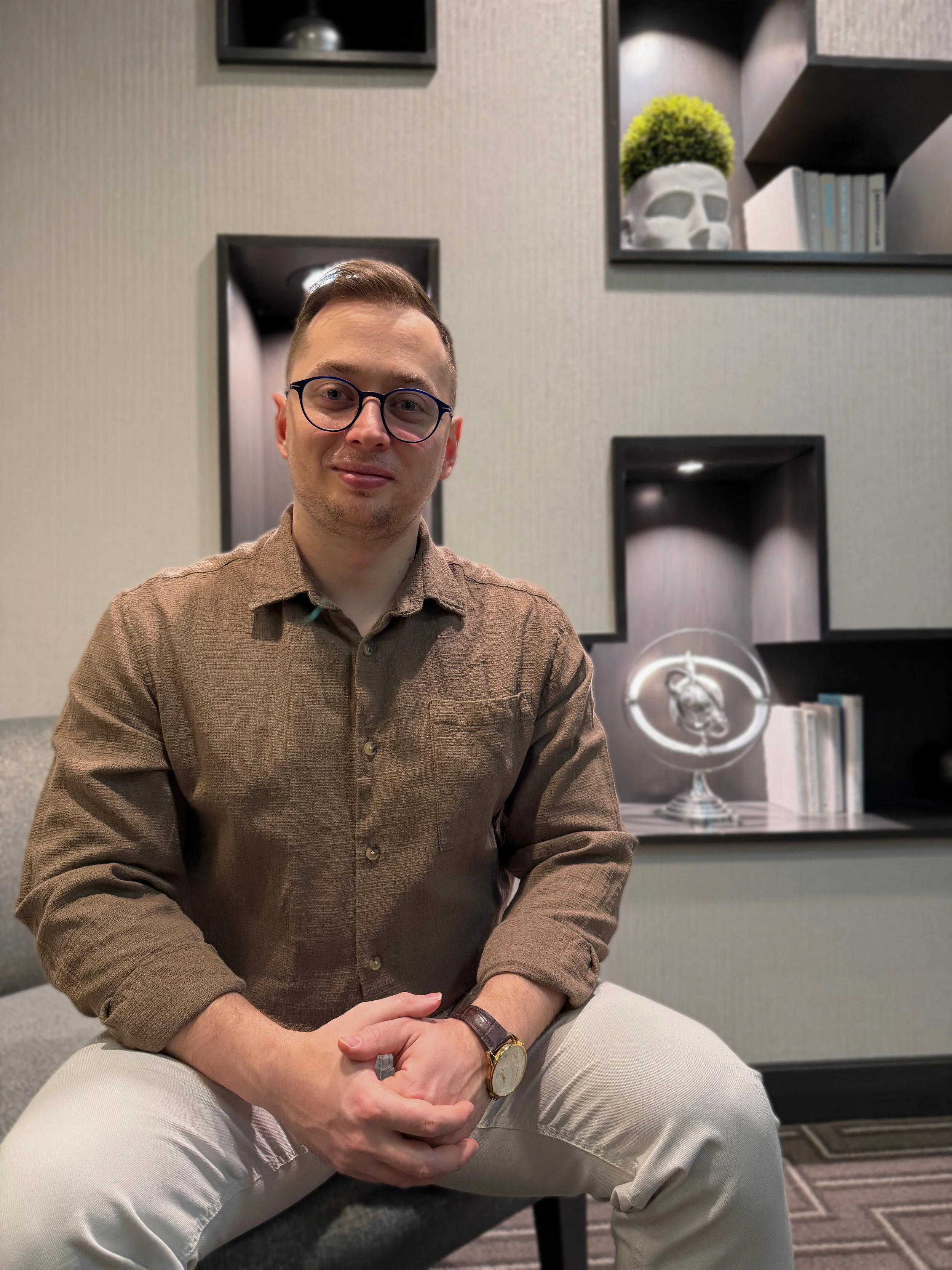 Kadir Iskesen sitting on a bench with his hands clasped, wearing a brown button-up shirt, beige pants, glasses, and a watch, in a modern indoor setting with decorative wall shelves, plants, books, and a globe.