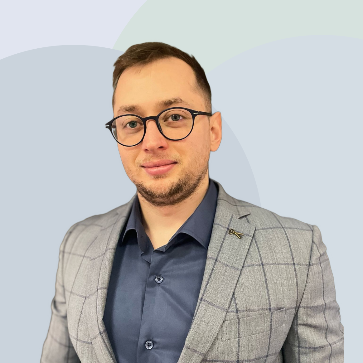Kadir Iskesen with glasses and a beard wearing a gray checkered blazer and a blue shirt, standing against a light background.