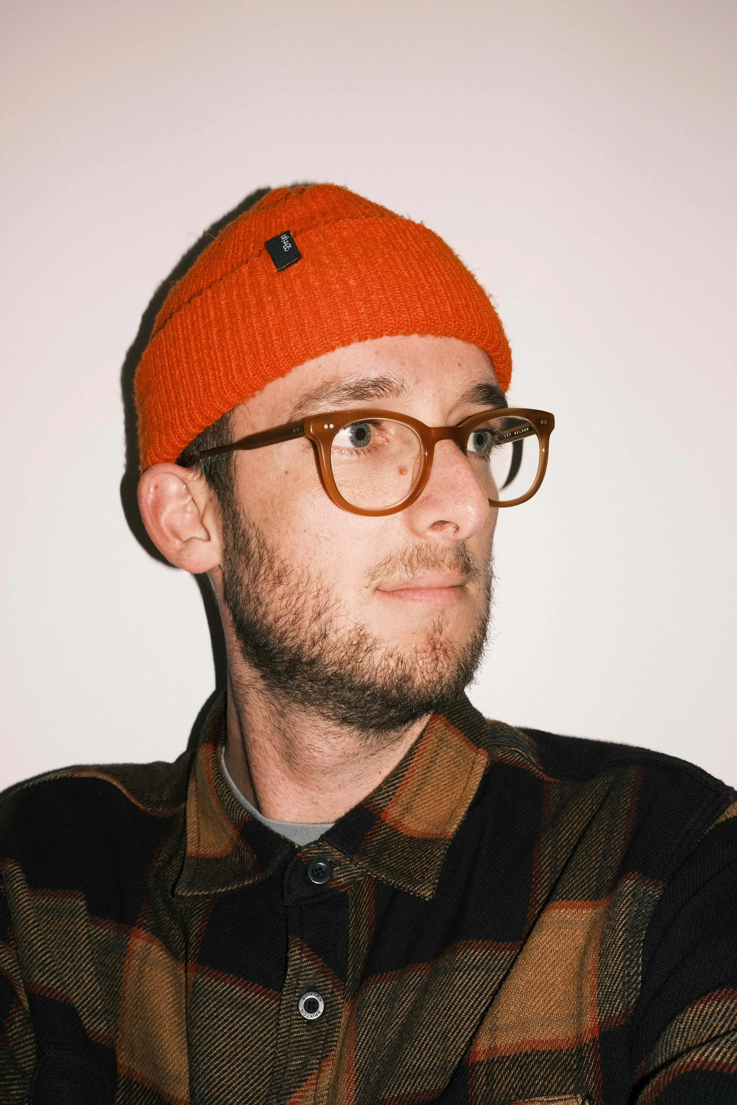 Close-up of a young man with light skin, brown hair, and facial hair wearing transparent brown glasses, an orange knit beanie, and a plaid shirt in shades of black, brown, and orange, against a plain light background.