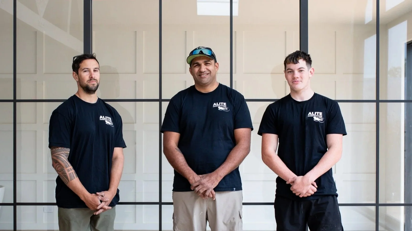 Three men in black T-shirts standing indoors in front of a glass wall.
