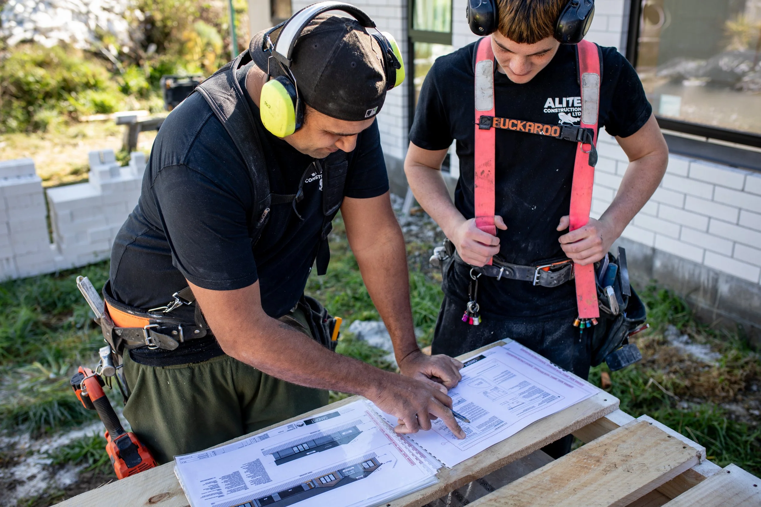 Two construction workers reviewing blueprints at a building site, wearing safety gear including ear protection and harnesses.