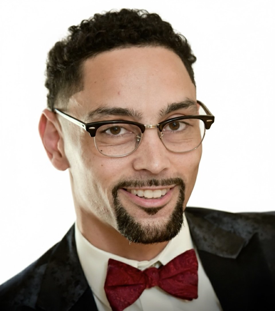 A man with curly dark hair, glasses, and a neatly trimmed goatee, smiling at the camera, dressed in a tuxedo with a red bow tie, against a plain white background.