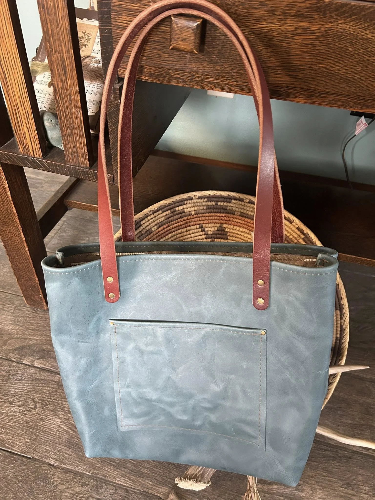 A light blue tote bag with brown leather straps hanging on a hook. The bag has a front pocket and is made of a textured material. It is placed on a woven chair with a wooden surface nearby.