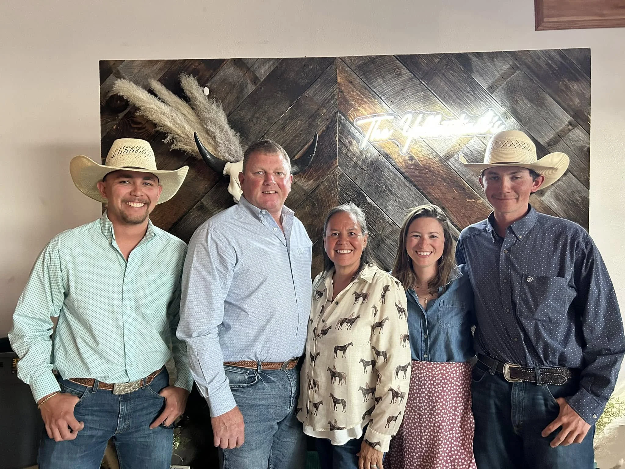 Five people posing in front of a wooden wall with a neon sign and deer skull with antlers. The group includes men and women wearing casual and western-style clothing, with two men wearing cowboy hats.
