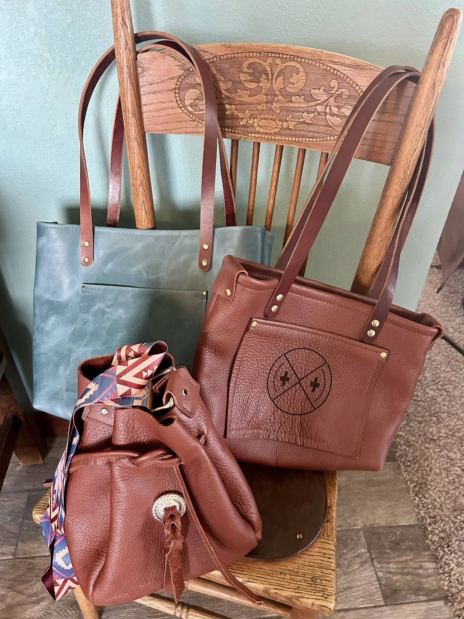 Three handbags on a wooden chair, with a light blue tote bag and two smaller leather bags in reddish-brown and pink, featuring a colorful scarf tied to one of the smaller bags.