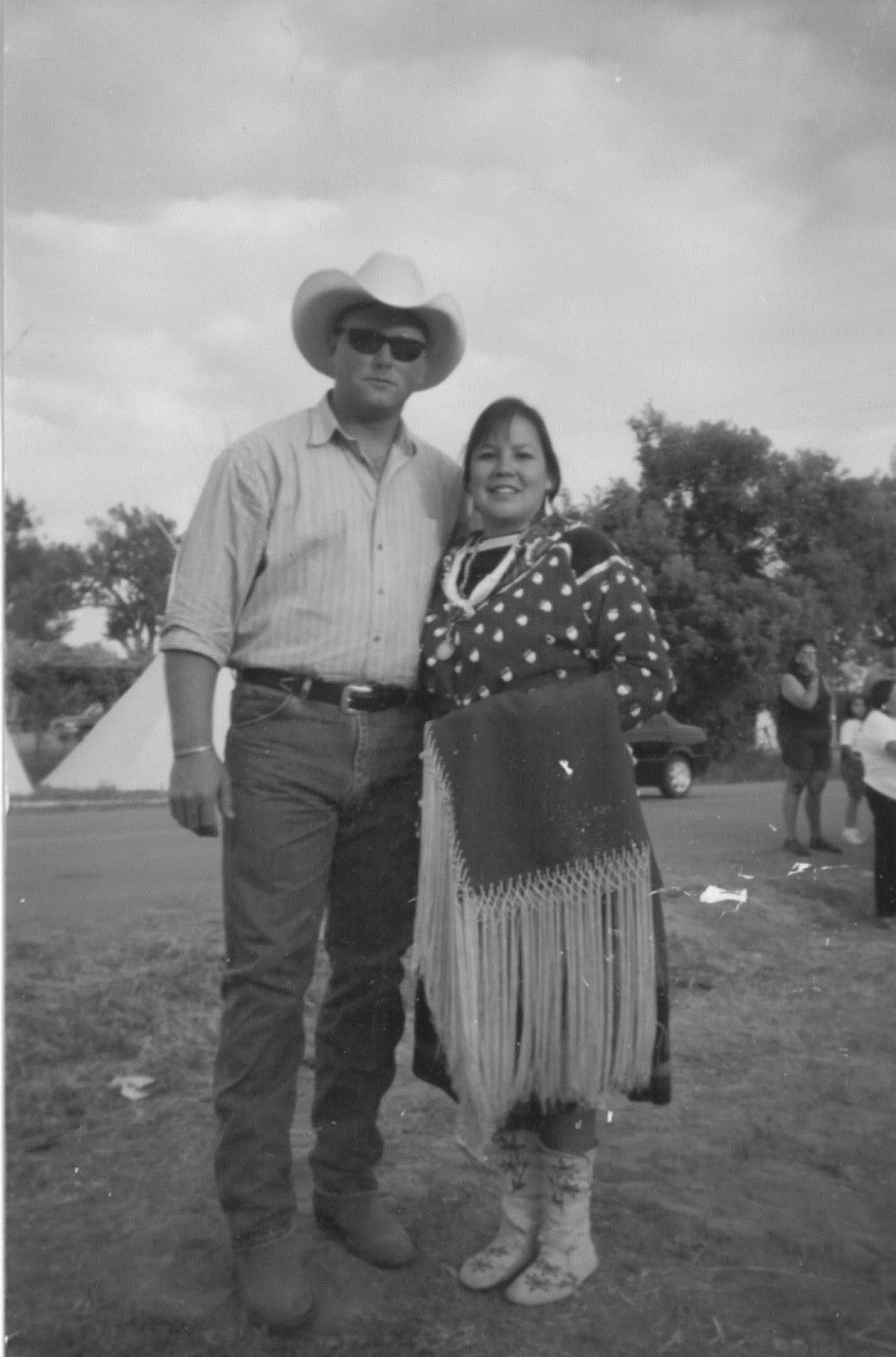 A man wearing a cowboy hat, sunglasses, a striped shirt, and jeans stands next to a woman in traditional Indigenous clothing with a beaded necklace and fringed skirt. They are outdoors with tents and trees in the background.