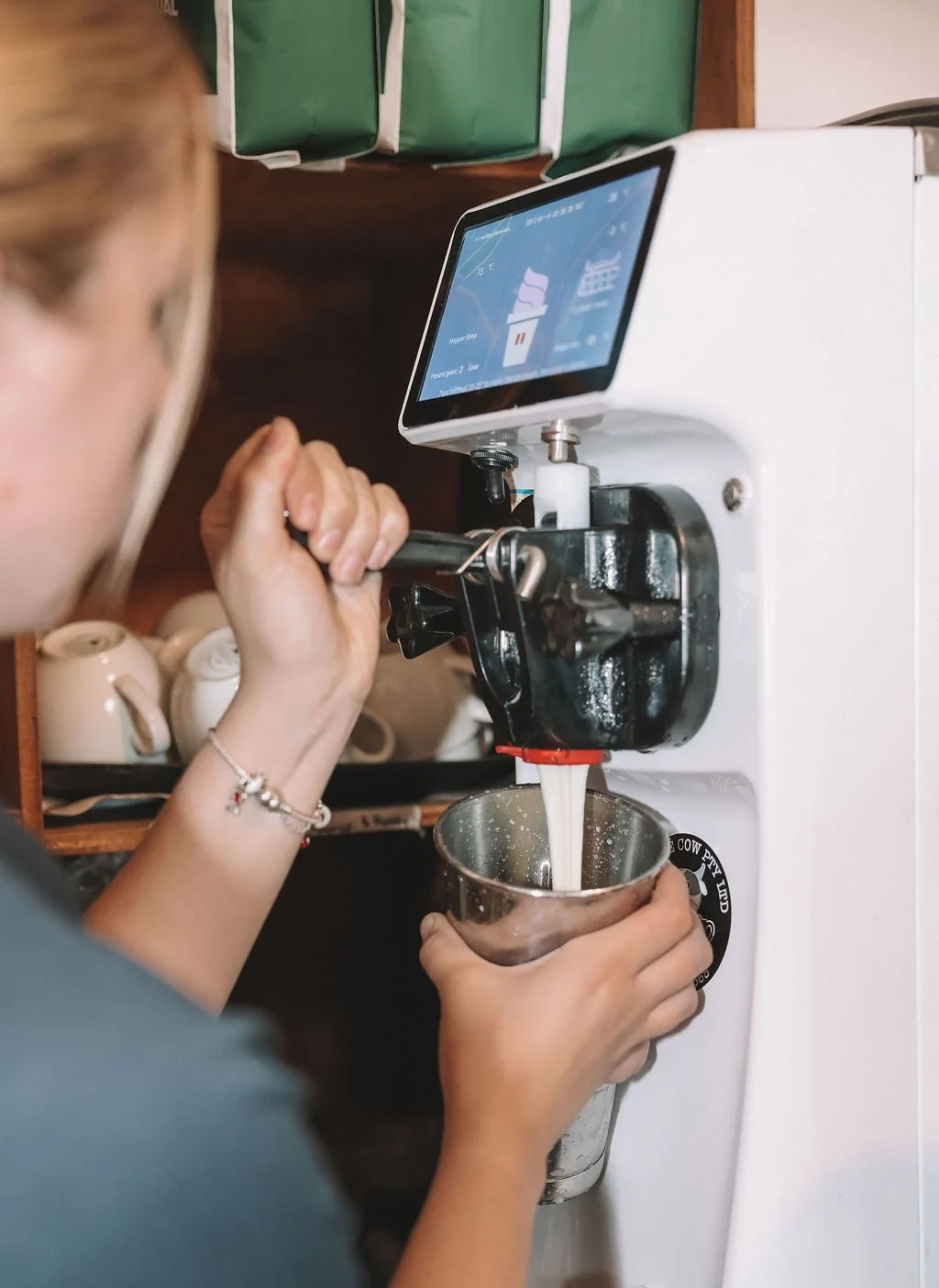 A person making ice cream using an ice cream machine with a digital display, pouring the soft serve into a metal cup.