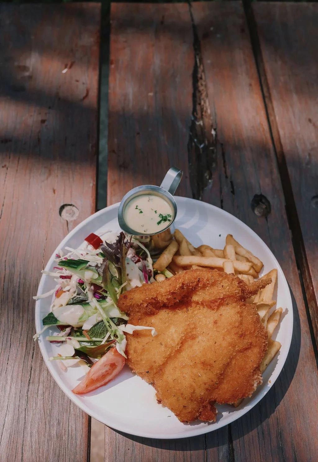 A plate with fried fish, French fries, a side salad, and tartar sauce on a wooden table.