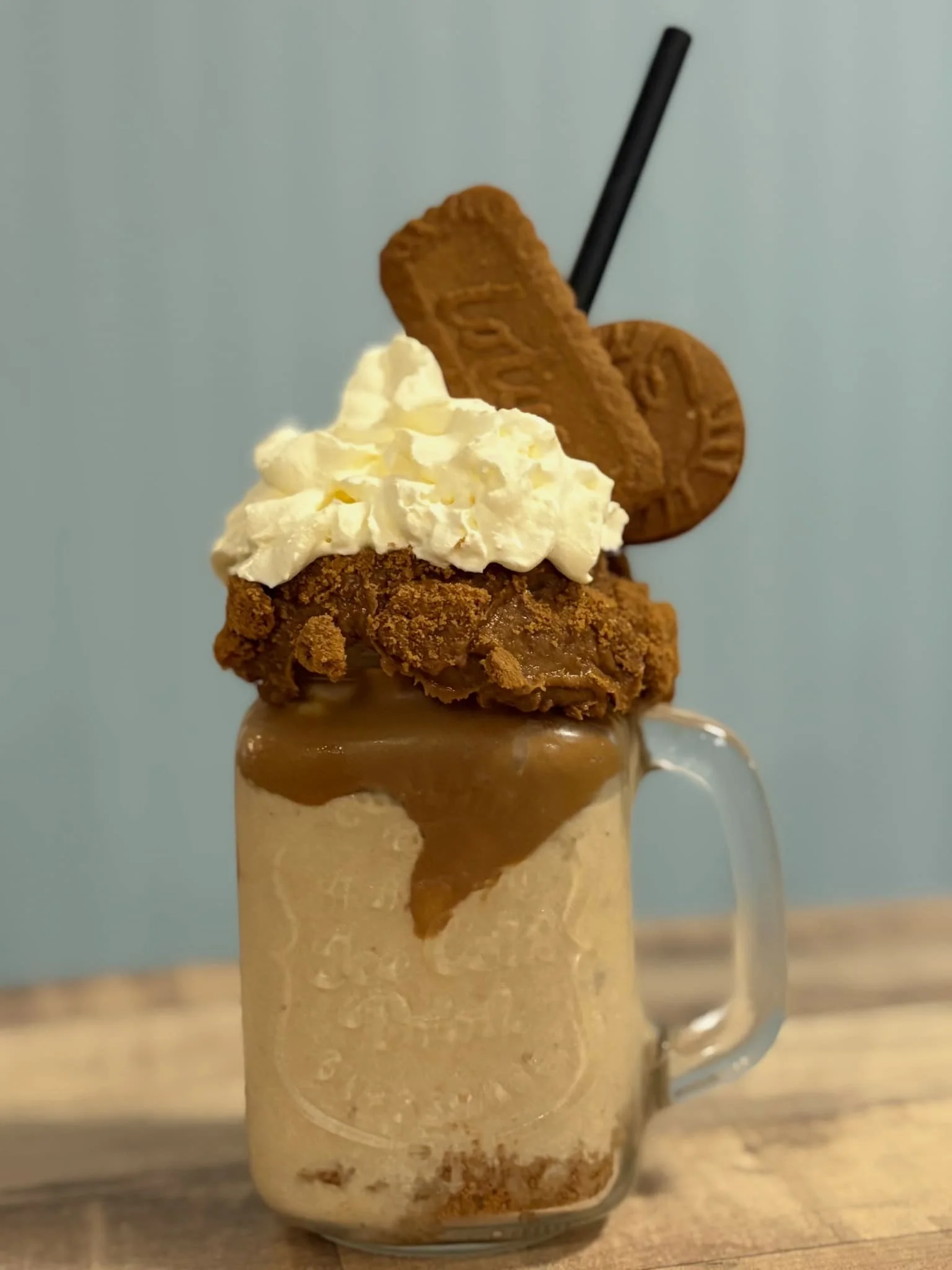 A layered dessert in a glass mug with whipped cream, caramel sauce, a hormone cookie, a vanilla cookie and a black straw on a wooden surface.