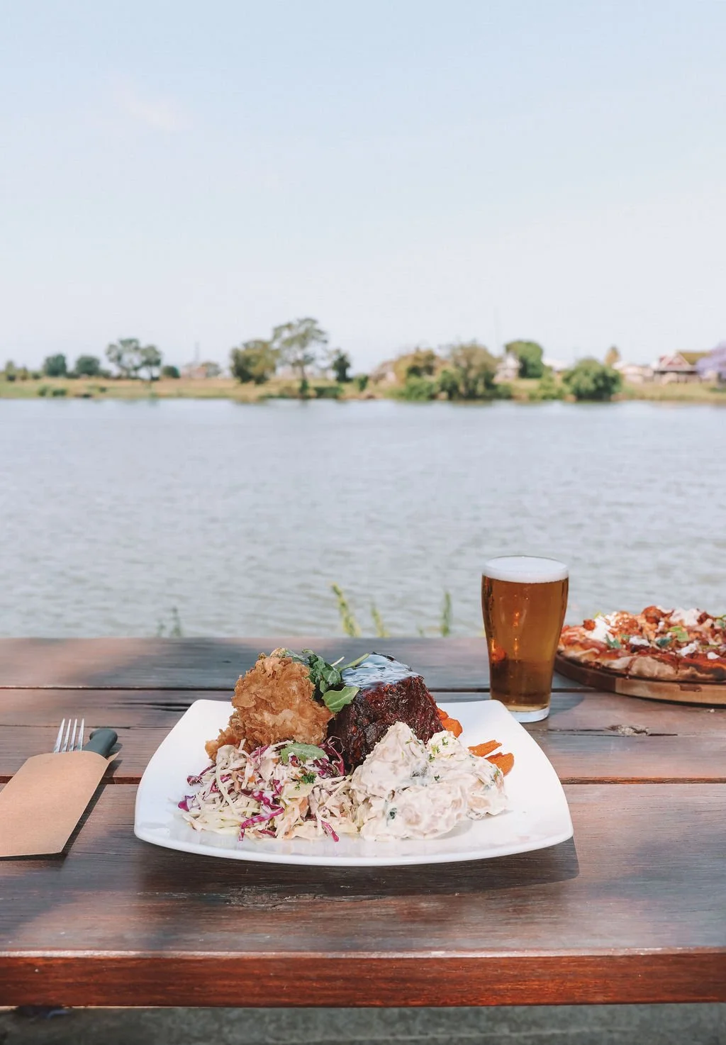 A plated meal with fried chicken, barbecue ribs, and coleslaw on a table overlooking a lake, with a glass of beer and a pizza in the background.