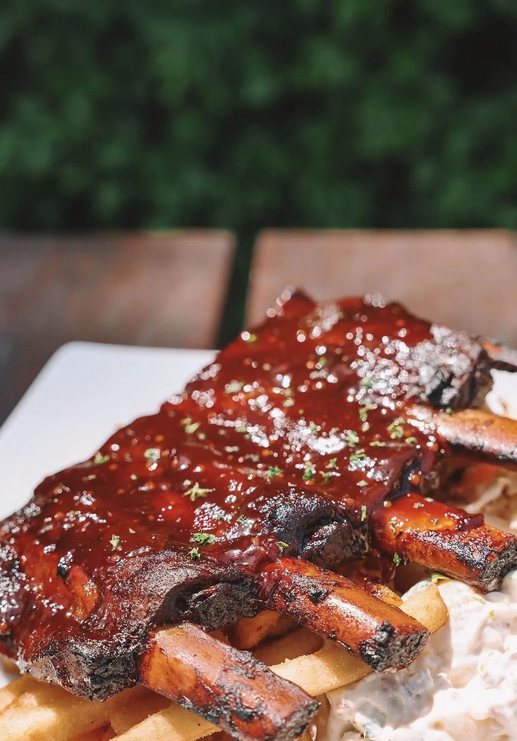Close-up of grilled ribs with barbecue sauce served on a white plate, with French fries and coleslaw underneath, blurred green background.