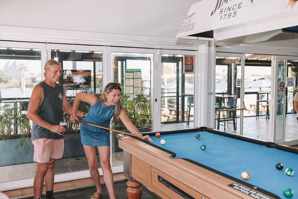 Two women playing pool at an indoor recreational venue, one woman is taking a shot while the other watches, holding a drink.