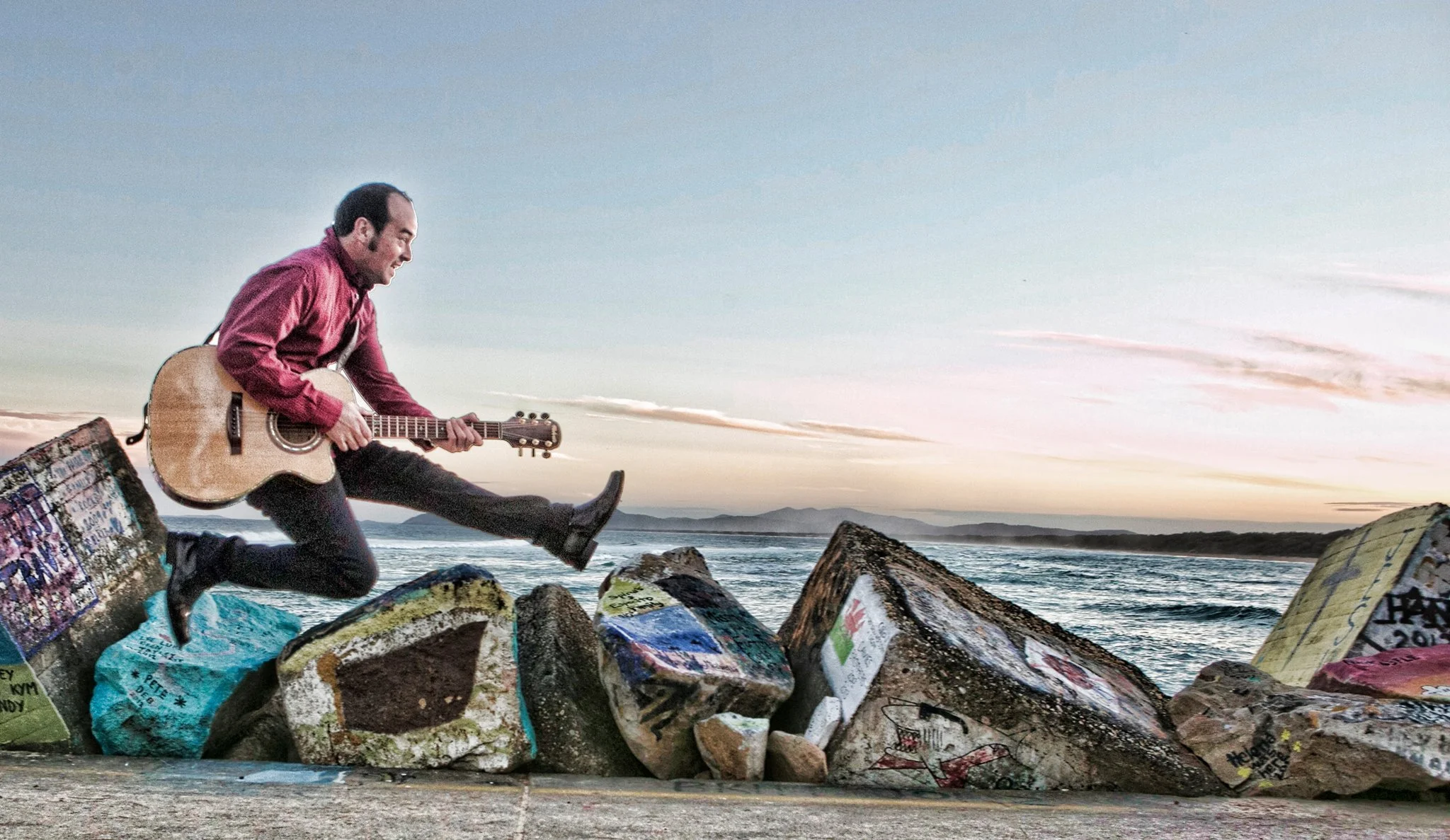A man in a red shirt and black pants playing an acoustic guitar while jumping on large painted rocks on a beach at sunset.
