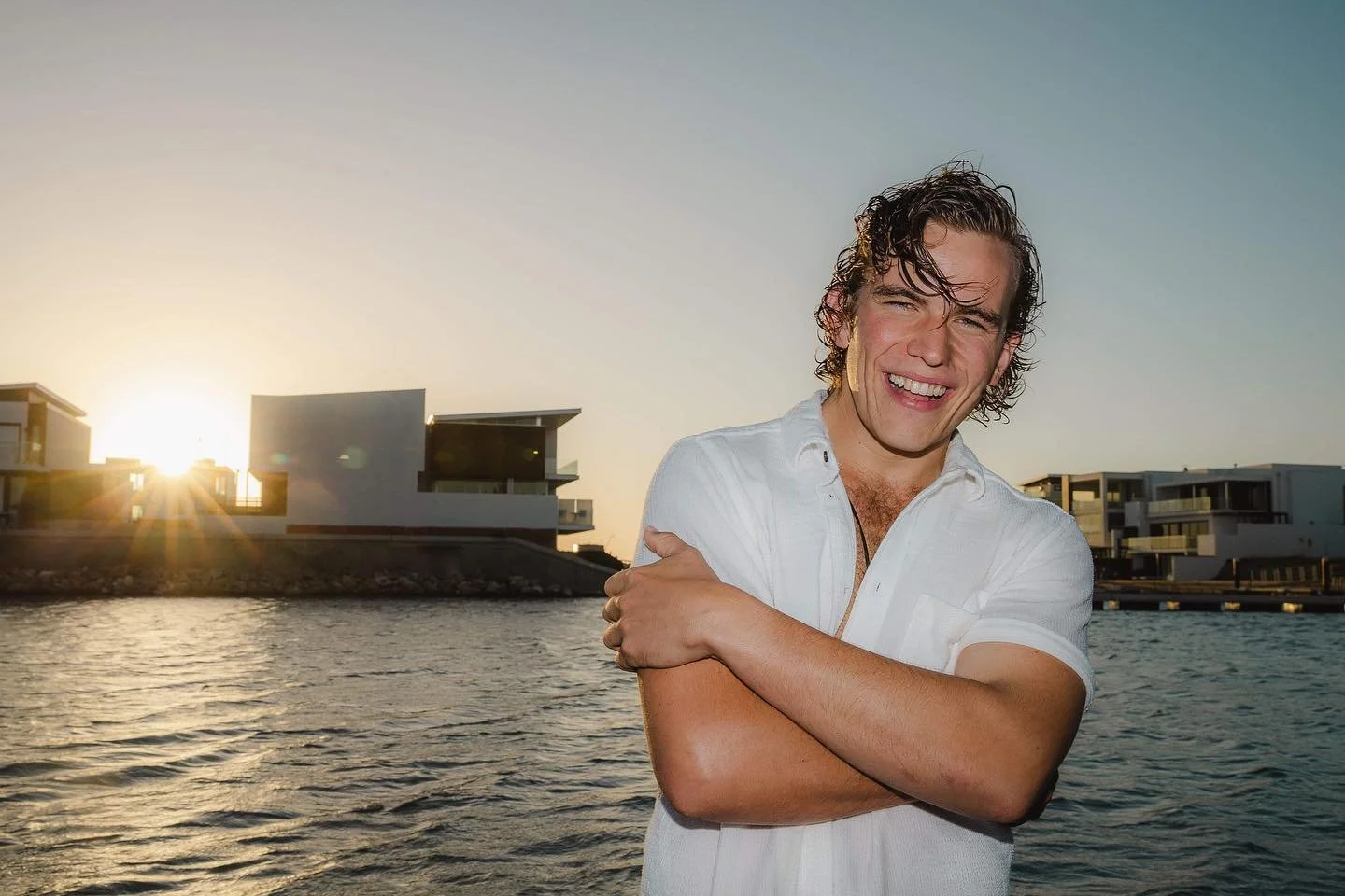 A young man with curly hair and a white shirt, smiling with arms crossed, standing near water at sunset with modern buildings in the background.