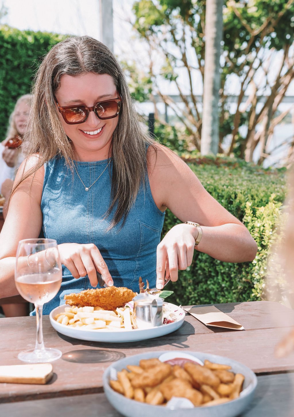 Woman wearing sunglasses and a blue sleeveless dress, smiling while cutting food at an outdoor dining table. There is a glass of rosé wine and a bowl of fried chicken on the table.