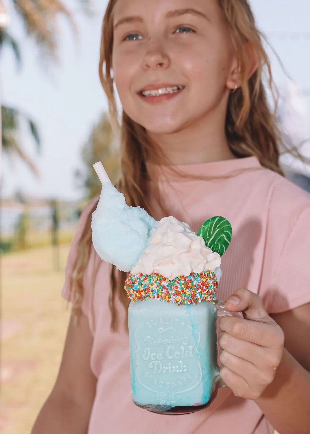 A woman in a pink shirt holding a large, colorful milkshake with whipped cream, rainbow sprinkles, cotton candy, a green leaf decoration, and a straw, outdoors with trees in the background.