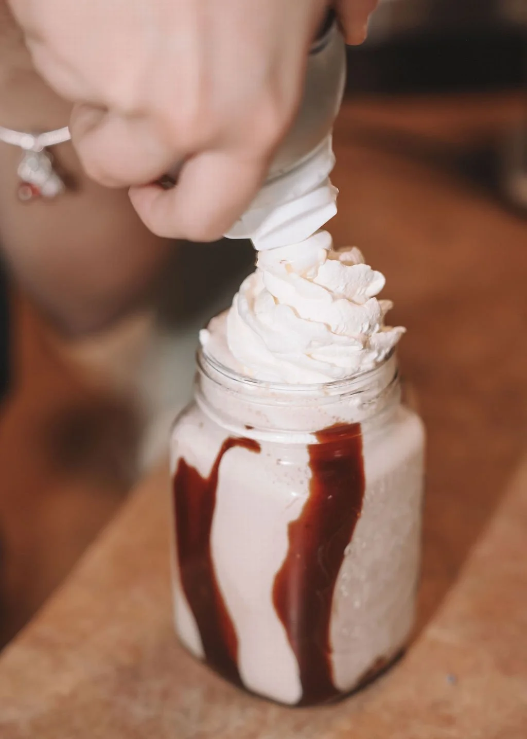 Person's hand squeezing whipped cream onto a chocolate milkshake in a jar with chocolate syrup drizzle.
