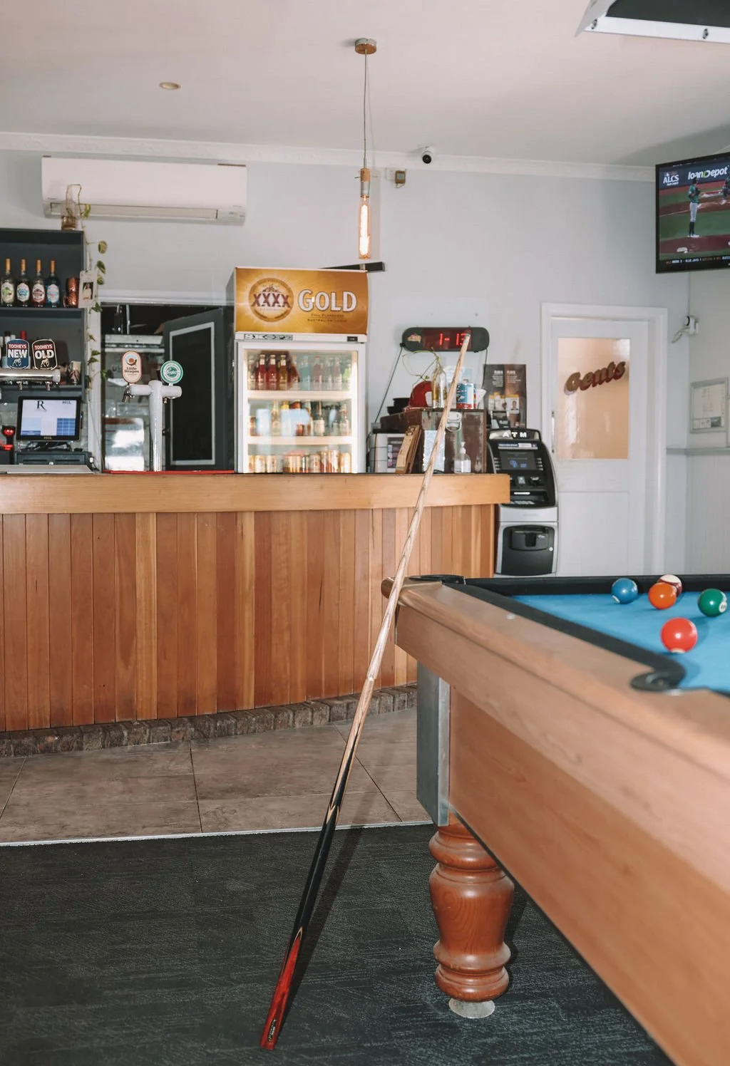 A pool table with a cue stick resting against the side, near a bar area with a refrigerator, a TV showing a baseball game, and various beverage bottles and paraphernalia.