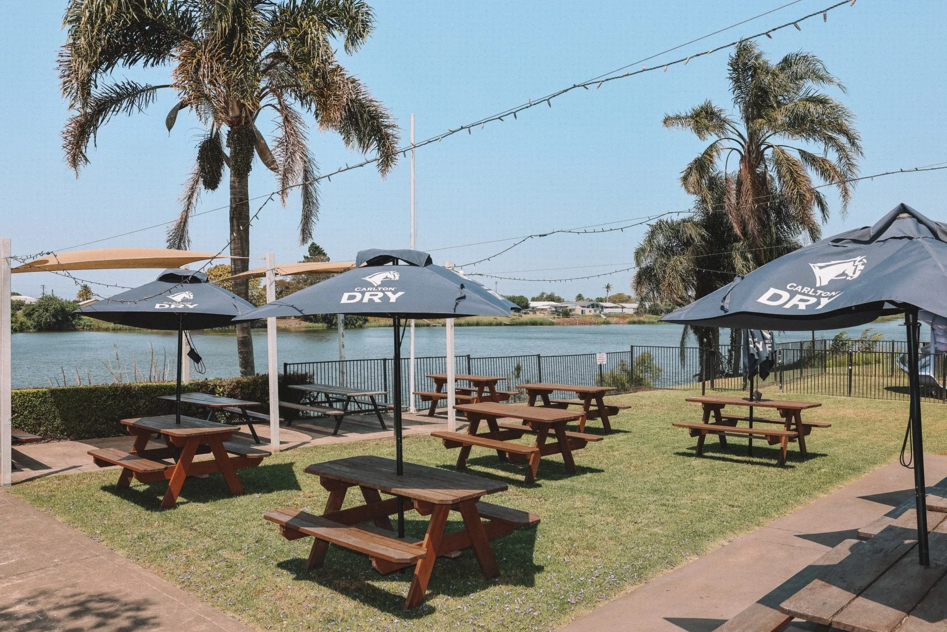 Outdoor seating area by the water with wooden picnic tables and large umbrellas, surrounded by palm trees and string lights, with houses and a bridge visible in the background.