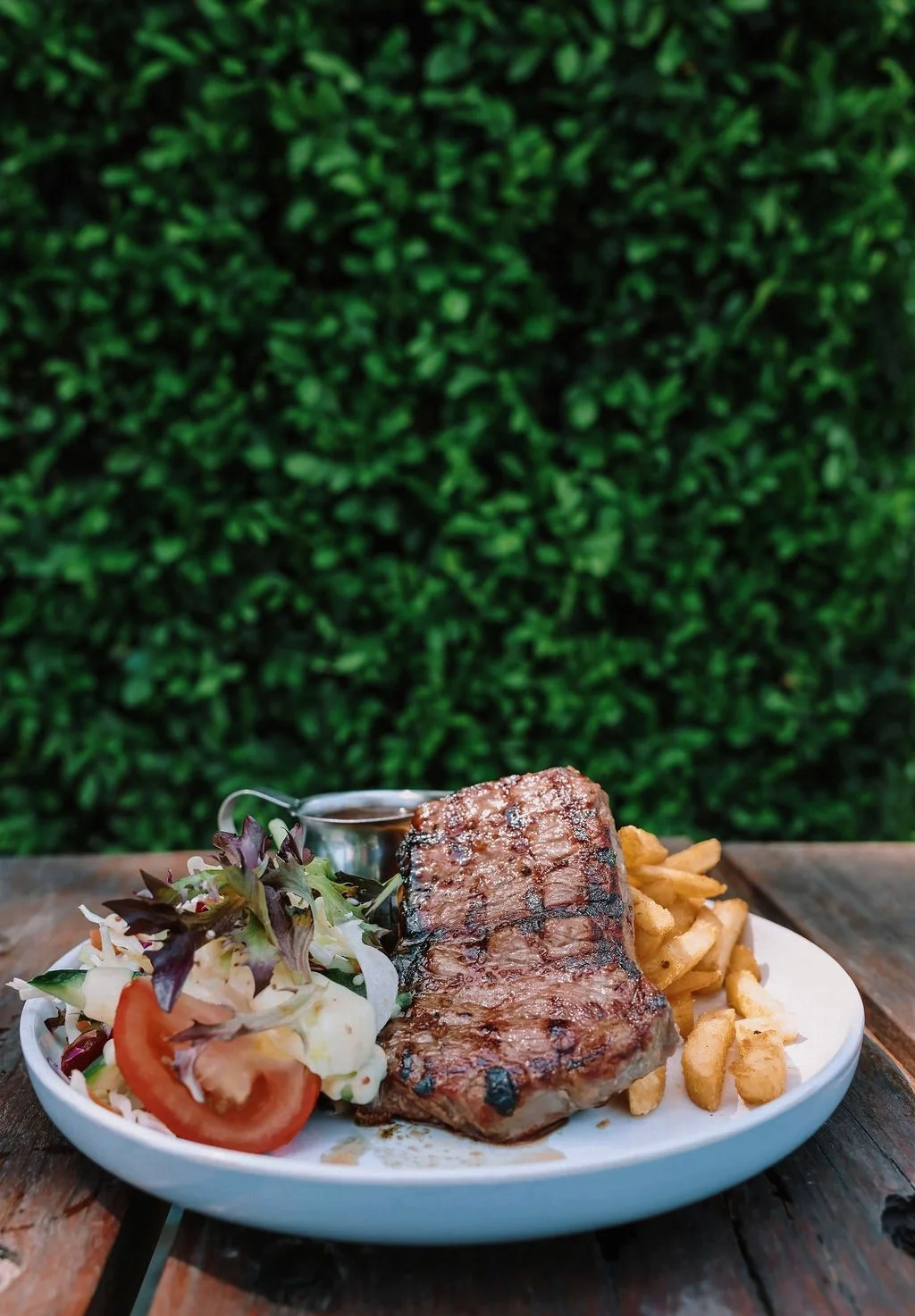 Steak with French fries, salad, and a small container of dipping sauce on a white plate outside.