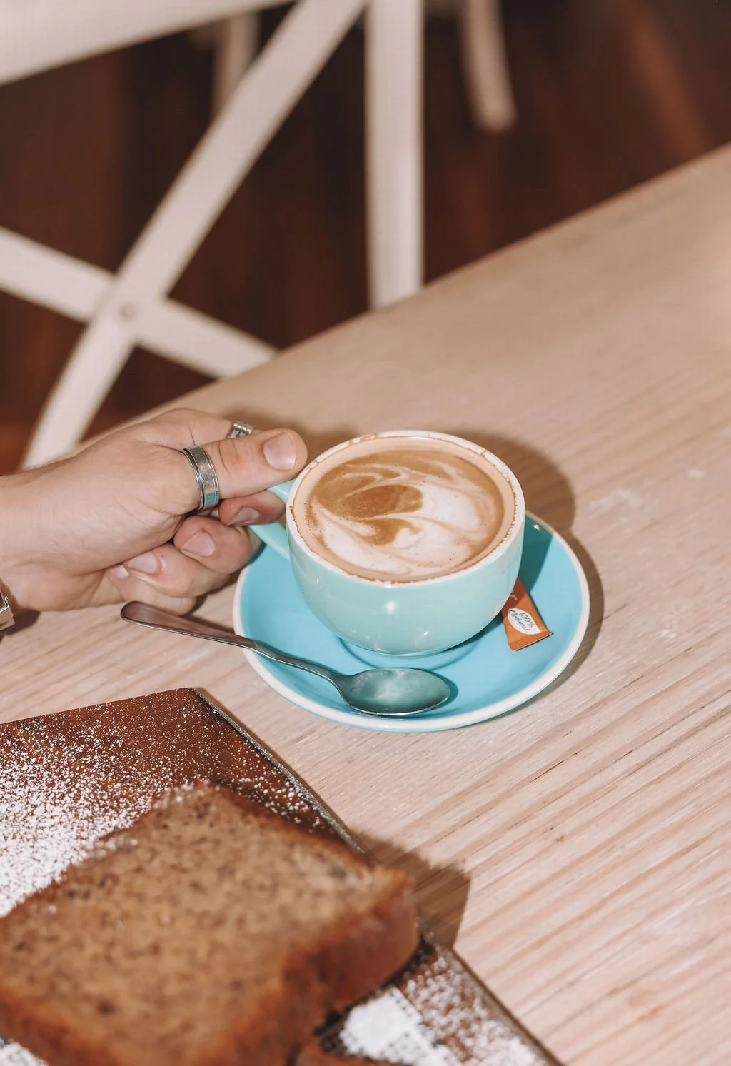 A hand holding a light blue cup of latte with foam art, placed on a matching saucer with a teaspoon, on a wooden table next to a plate of toast with powdered sugar.