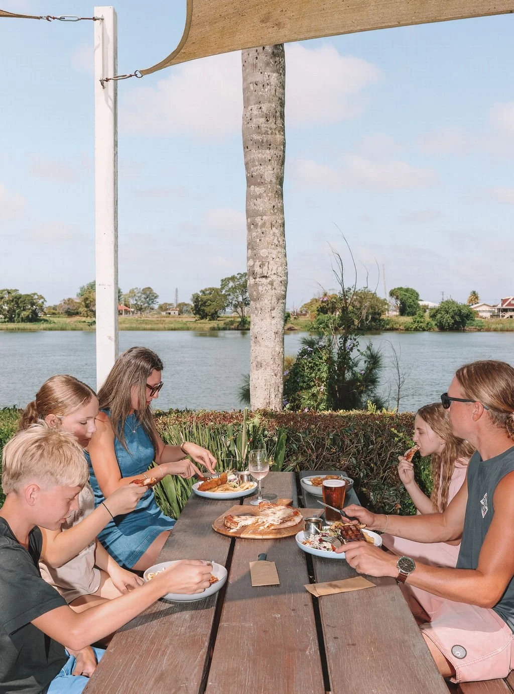 A family of five eating outdoors at a picnic table by a river, with trees and houses in the background.