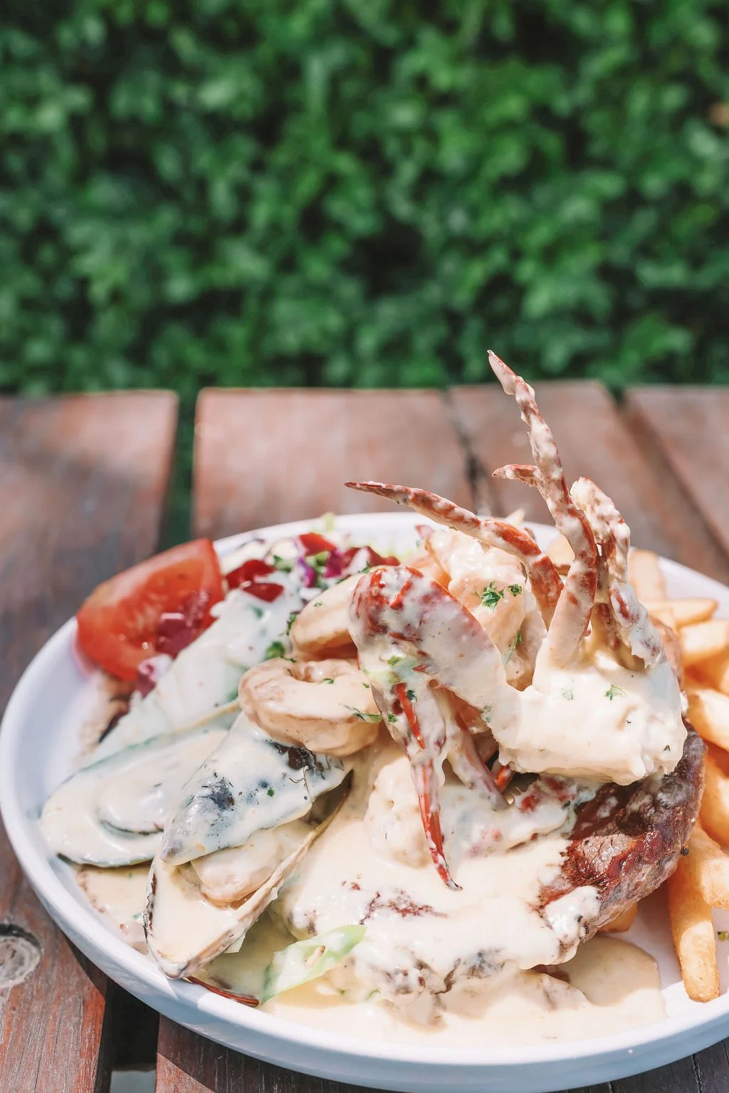 Seafood platter with shrimp, mussels, crab legs covered in creamy white sauce, served with sliced tomatoes and French fries on a white plate outdoors.