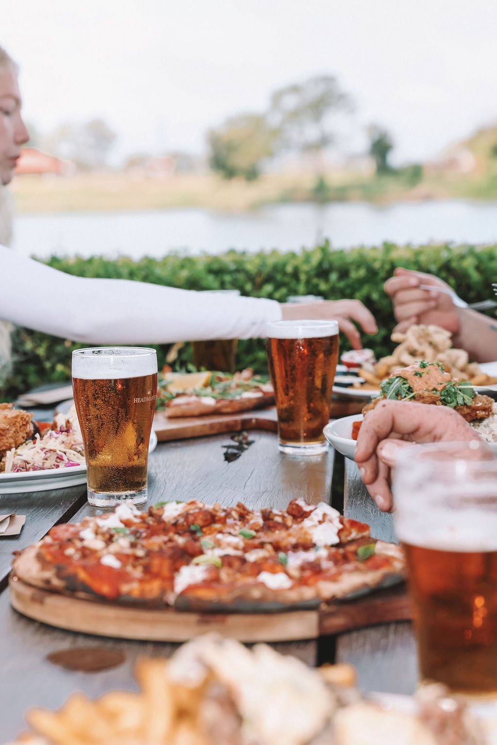 People enjoying a meal outdoors with pizza and beer, overlooking a body of water and greenery.