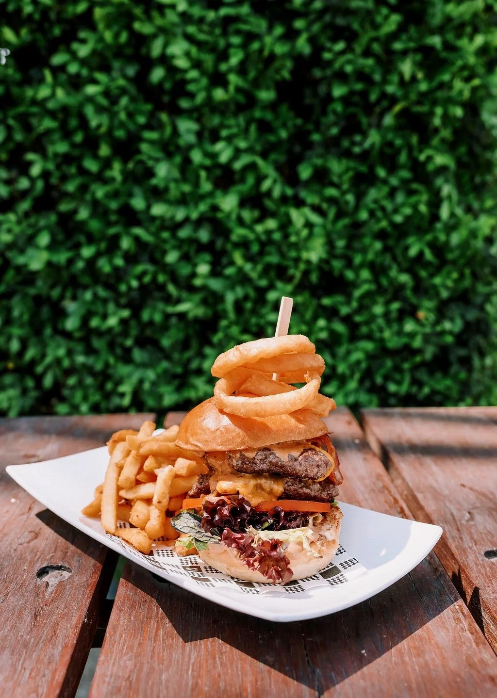 A burger with onion rings on top, French fries on the side, served on a white plate on a wooden table with a green leafy background.
