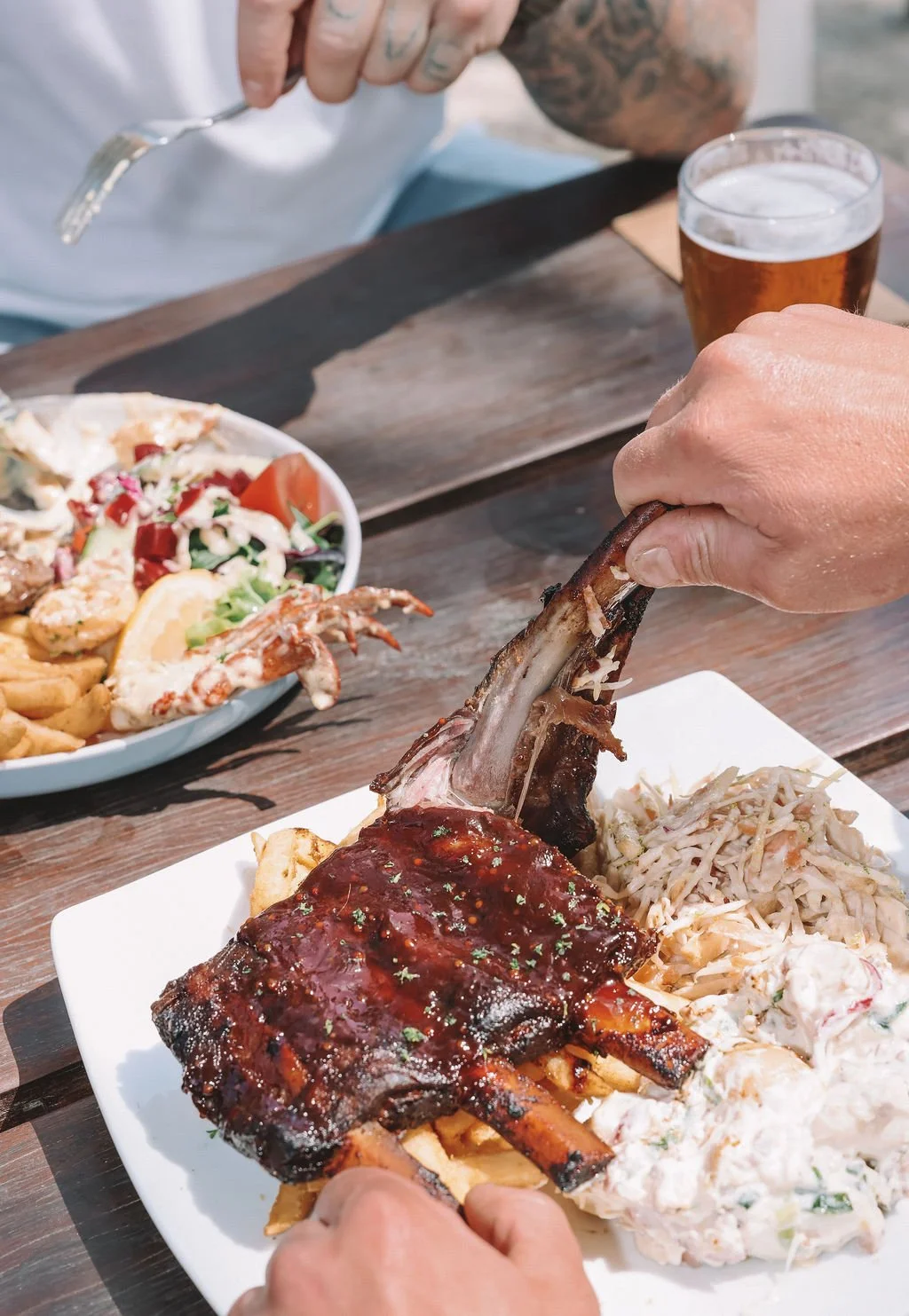 A person is holding a cooked beef rib by the bone over a white plate with side dishes including coleslaw, fries, and barbecue sauce. There is a bowl of salad and a glass of beer on the table in the background.