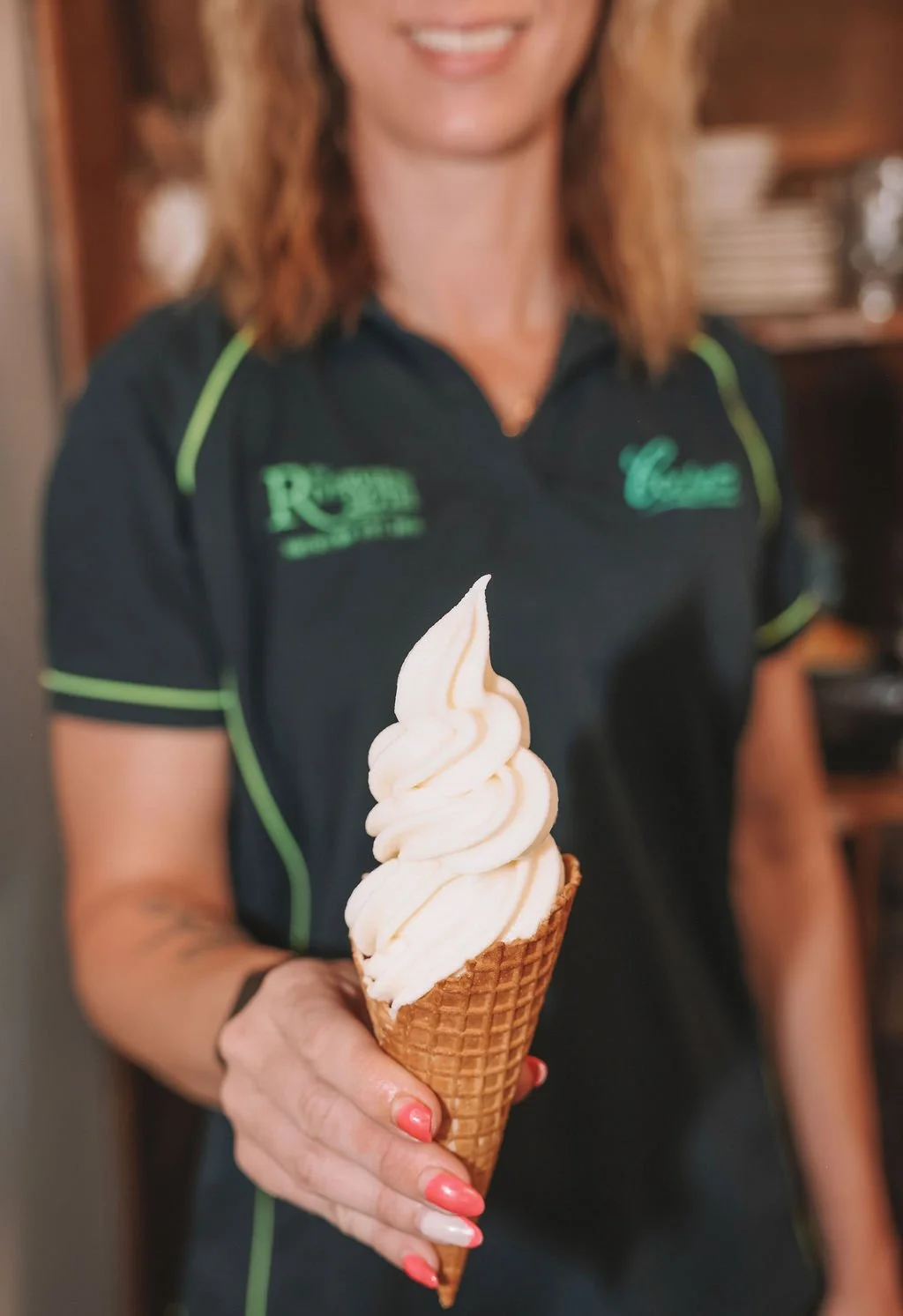 A woman in a black polo shirt with green accents holding a vanilla soft serve ice cream cone.
