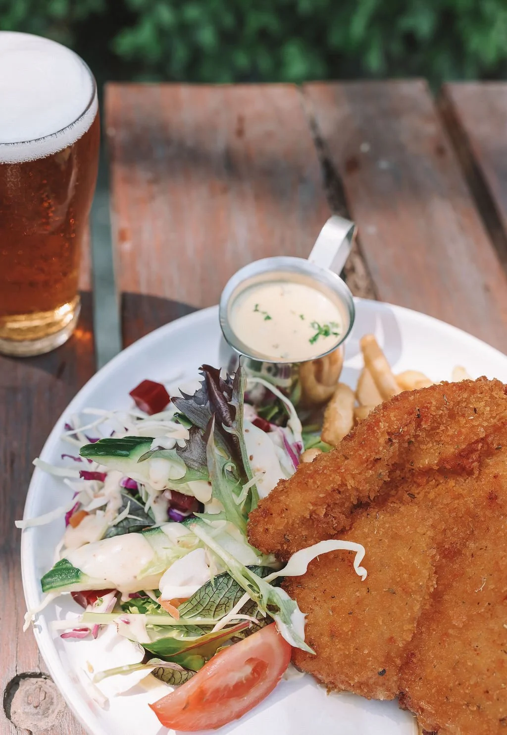 Fried fish fillet with salad, French fries, and creamy sauce on a white plate, accompanied by a glass of beer.