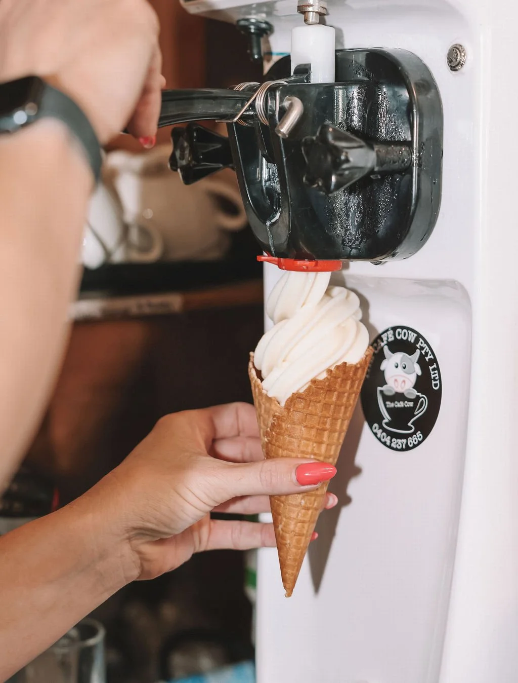 Person holding a vanilla ice cream cone under a black soft serve ice cream machine.