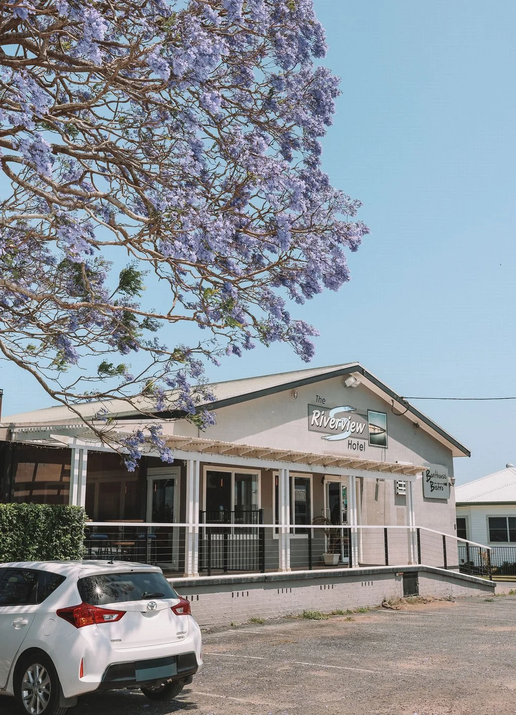 Exterior of The Riverview Hotel with a large tree with purple flowers in the foreground and a parking lot with a white car in front.