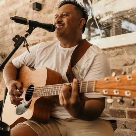 Man singing and playing acoustic guitar at a microphone in a rustic setting.
