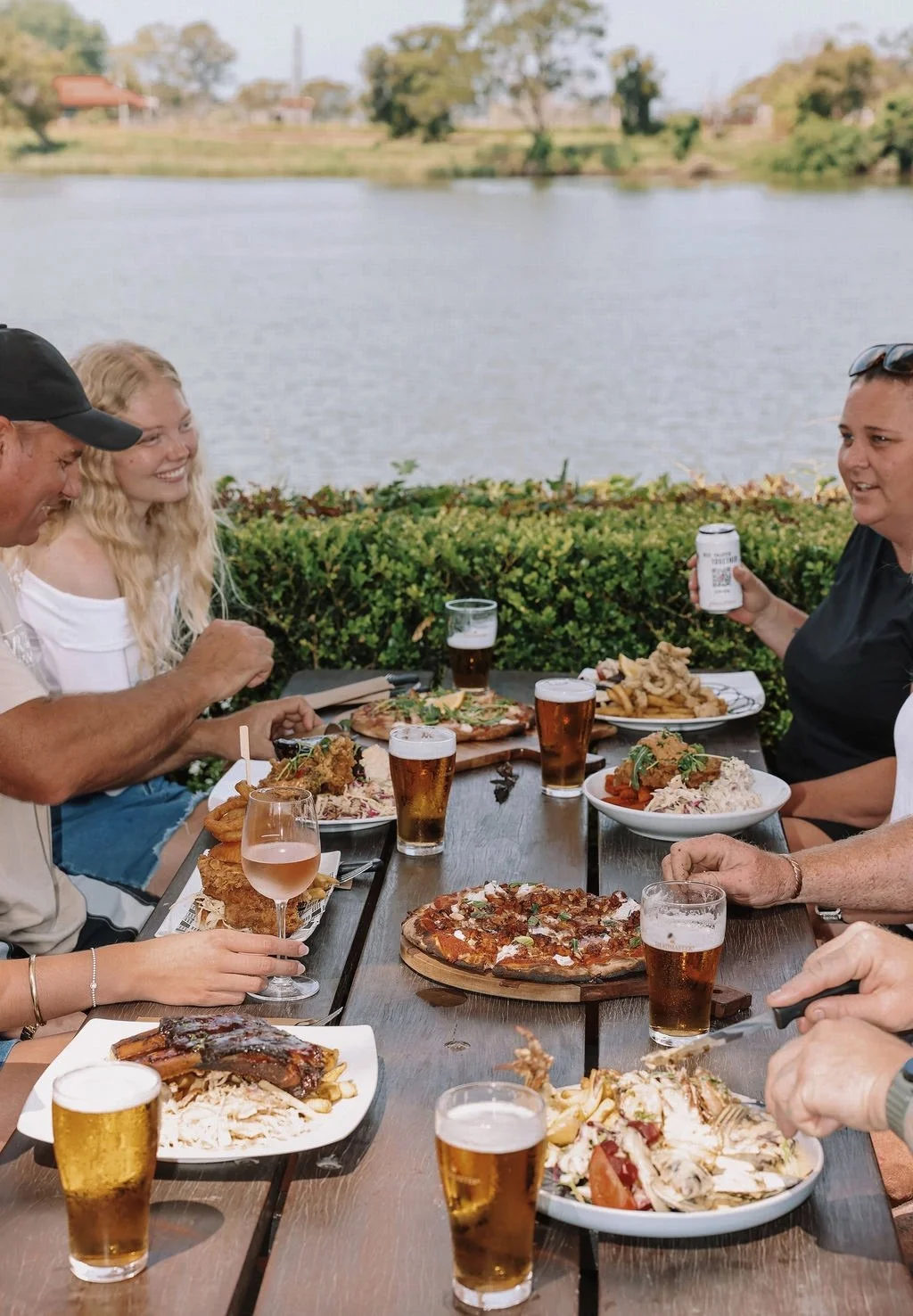 People enjoying outdoor dining at a lakeside restaurant with pizza, fried chicken, pasta, and drinks.